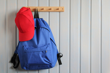 blue backpack and a red baseball cap hang on a wooden hanger on a white board wall. copy the space.