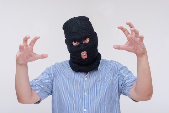 A scary and unhinged evil man wearing a black ski mask makes a threatening though comical gesture, showing his claws. Isolated on a white backdrop.