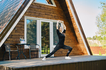 Sporty woman in sport clothing does stretching exercise on the terrace of wooden cabin - obrazy, fototapety, plakaty
