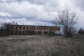 Fototapeta na wymiar A dilapidated building with broken windows amidst overgrown grass under a cloudy sky.