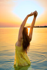 A young charming woman in a yellow dress stands on the seashore in front of a bright orange sunset.