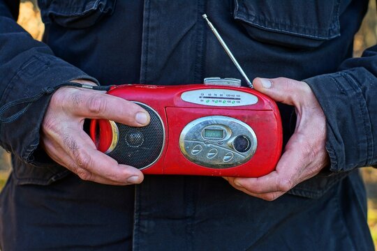 one old small plastic red, battery-powered radio with antenna and speaker, in the hands of a man dressed in a black jacket on the street during the day