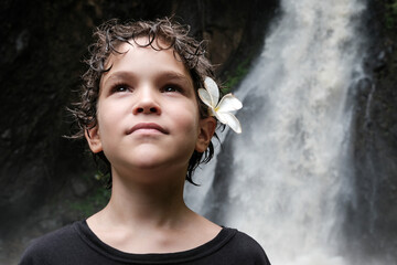 Portrait of a Caucasian boy with plumeria (frangipani) flower in his hairs on the background of a waterfall. Bali, Indonesia.