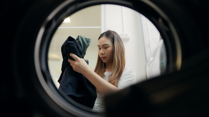 Young woman putting clothes in washing machine and sitting on floor in home laundry. She loads the washer with dirty laundry.