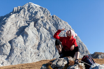 Adult Woman Hiker Hydrating with Water in Squatting Position on High Mountains Background