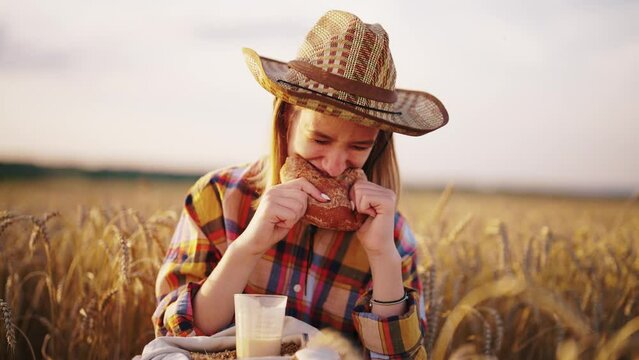 Young girl farmer in sun hat eats loaf of bread biting it drinking milk sits on wheat field with grain bag on knees. Harvesting, summer on farm, food business, agribusiness, woman worker has break.