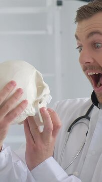 Insanely cheerful young attractive doctor holding a human skull skeleton in his hands. Vertical video