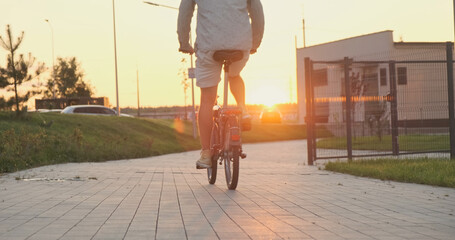 Man casual wear rides folding bicycle, sidewalk, back view, sunset, sky, urban, cars