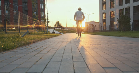 Man rides folding bicycle on sidewalk among high-rise apartment buildings to meet the setting sun, back view