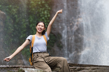 Fototapeta na wymiar Cheerful young woman with backpack sitting near waterfall and enjoying the splashing nature power. Traveling and freedom concept.
