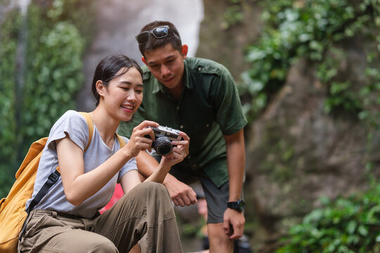 Happy young couple sitting at tropical waterfalls and checking pictures on camera. Vacation and traveling lifestyle concept.