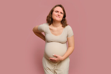 A pregnant woman holds her aching back against a studio pink background. Pregnancy in a woman with a belly