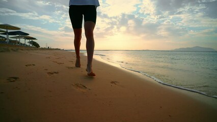 Barefooted woman jogging along sandy sea ocean beach holding bottle of water at sunset. Outdoors training, running, wellness sport training, workout of female. Active lifestyle, body care concept.