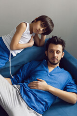 Obraz premium Portrait of a family father and son sitting on a blue sofa in the studio opposite the window. A hipster man with a beard and blue eyes in a shirt hugs his child.