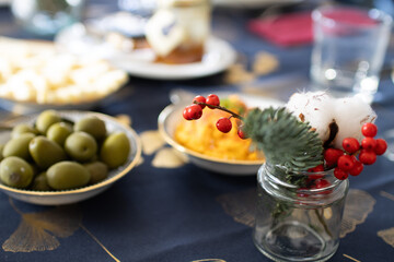 A festive table setting with a cotton branch, olives, olive oil, and a glistening dish of hummus on a blue gold-patterned cloth. Christmas table setup with mediterranean food