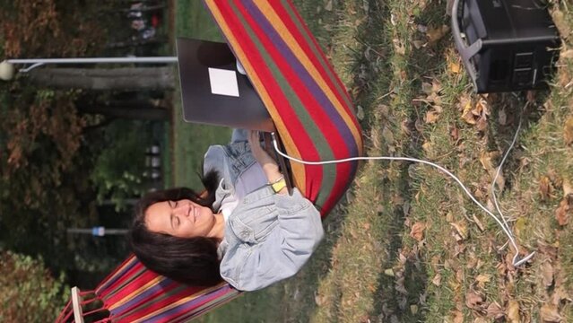 A young woman in a hammock in a summer garden is working on a laptop. Charging a laptop from a portable power station