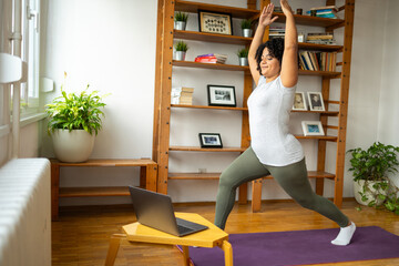 Happy woman practicing warrior pose during online classes on laptop in yoga room