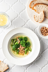 Vegetarian minestrone soup with green vegetables and beans served in white plate on white tile background, top view