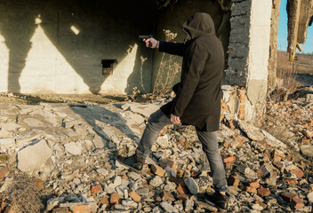 Man armed with a gun. A man dressed in a black coat with a hood commits a crime in the ruins of an...
