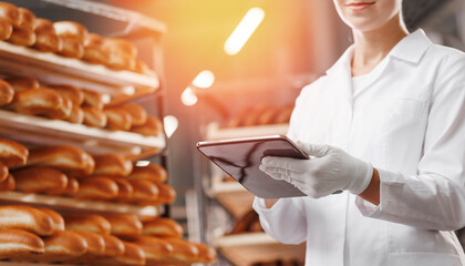 Baker woman in chef uniform use tablet computer for control quality of craft bread in bakery factory, sun light. Banner modern food industry