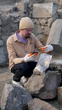 A male archaeologist with glasses and a beard examines pieces of ceramic pasuda shards found during archaeological excavations and uses a smartphone to compare relics. Vertical screen