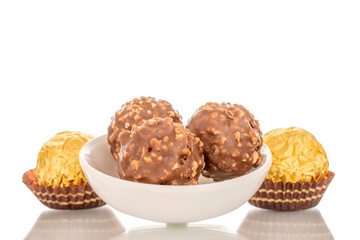 Several chocolate candies with nuts in a paper wrapper with a white ceramic saucer, macro, isolated on a white background.