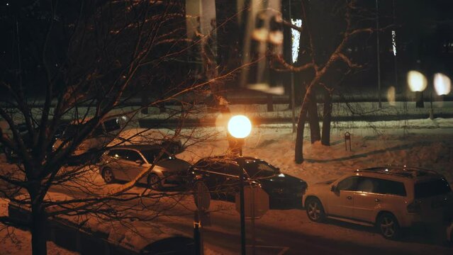 View from the window into the courtyard, where cars stand in the light of lanterns and the road along which cars are driving on a winter evening.