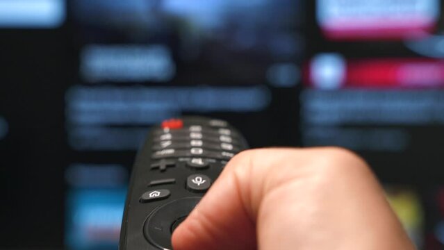 Close-up of a smart TV remote control and man's hand scrolling and choosing content for watch on online video platform. Blurred television screen in the background