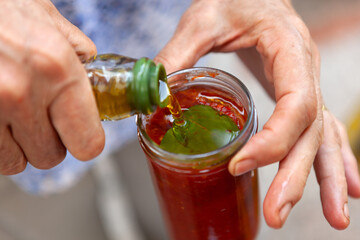Woman hands completing a homemade tomato sauce in a glass jar, adding fresh basil leaves and olive...