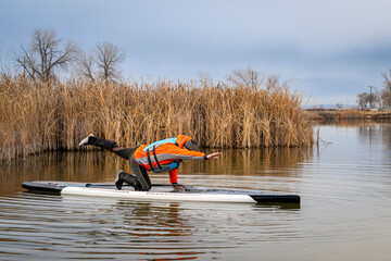 senior male stand up paddler is practicing yoga poses on his paddleboard on a lake in Colorado,...