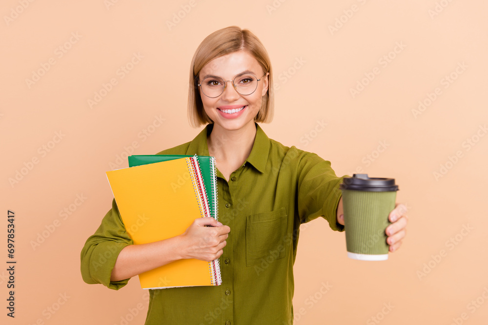 Sticker Photo of friendly cheerful woman in glasses hold copybooks give you paper cup of hot cappuccino isolated on beige color background