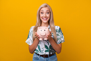 Fototapeta na wymiar Photo of impressed ecstatic cute girl with long hairdo dressed print shirt hands hold piggy box isolated on yellow color background