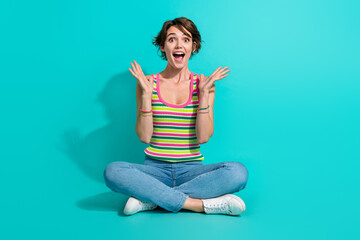 Full size photo of astonished positive girl sit floor raise hands cant believe isolated on teal color background