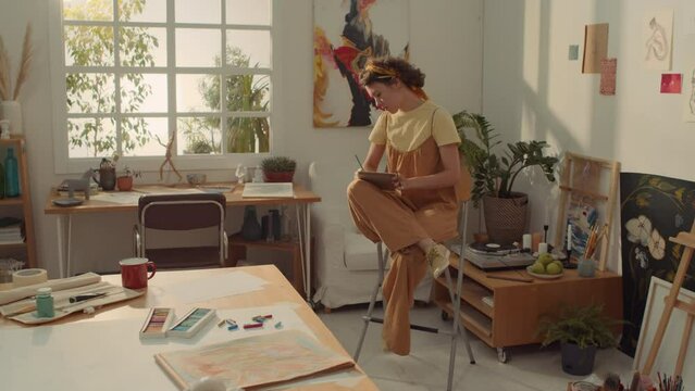Full shot of busy young Caucasian female artist wearing jumpsuit and t-shirt sitting on high chair, drawing sketch on notepad in pencil while working in studio, looking at window