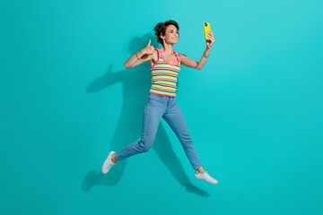 Fototapeta na wymiar Full size photo of charming sportive girl jump hold smart phone take selfie show thumb up isolated on teal color background