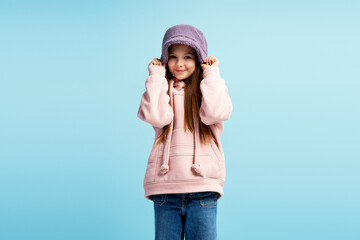 Smiling little fashion model wearing stylish hat, looking at camera, posing on blue background