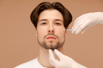 Handsome bearded young man getting under eye injections. Isolated on beige background. Beauty care,...