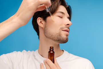 Close up young man applying serum on his face with pipette. Isolated on blue background. Daily beauty treatment concept
