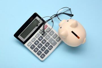 Calculator, glasses and piggy bank on light blue background, flat lay