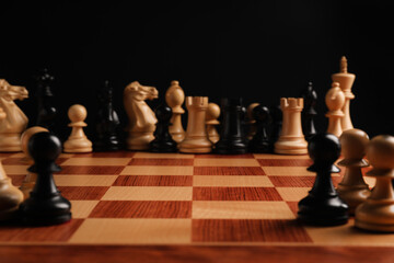 Many chess pieces on wooden checkerboard against black background, selective focus