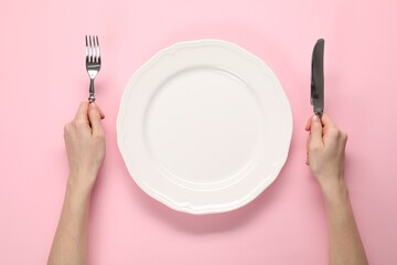 Woman with fork, knife and empty plate at pink table, top view