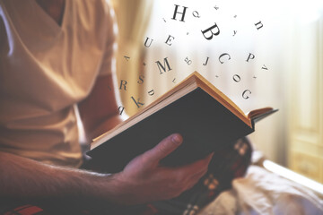 Young man reading book with letters flying over it on bed at home, closeup