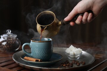 Turkish coffee. Woman pouring brewed beverage from cezve into cup at wooden table, closeup