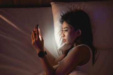 Asian woman using mobile phone smartphone laying on the bed in the bedroom. Sleepy exhausted, can...