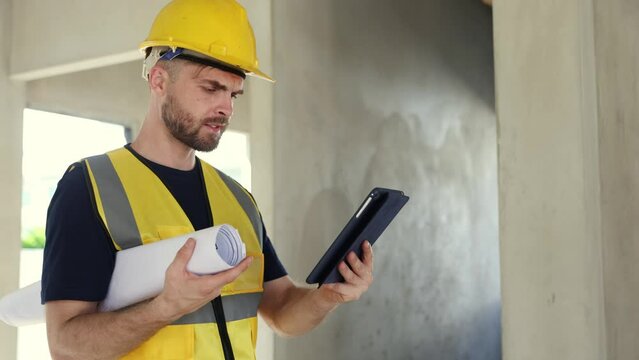 Male architect foreman watches progress by looking at construction blueprints holds laptop looks at interior design and details under construction in housing project looking at camera gives thumbs up.