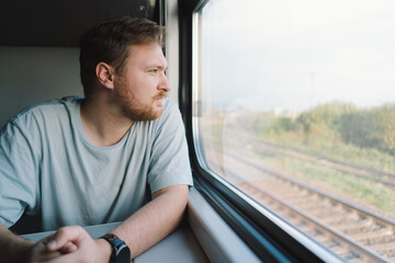 A man with a beard and mustache in a blue t-shirt while traveling by Railway train, sitting in the train and looking out the window. - obrazy, fototapety, plakaty
