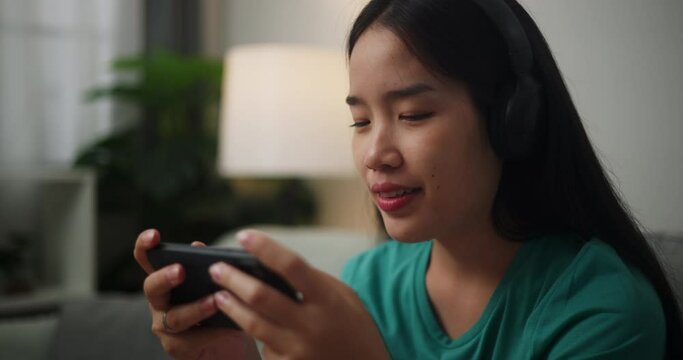 Footage of Young Asian woman wearing headphones enjoys playing online esport games on smartphone sitting on sofa in the living room at home,Gamer lifestyle concept.