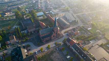 The image captures the Sint Jozef Church in Rijkevorsel from an aerial viewpoint at dawn. The rising sun casts a warm light on the church's brick architecture, illuminating the building and its