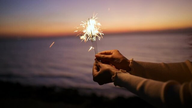 Closeup of hands holding sparklers, happy friends with fireworks against background of evening sunset sky on the beach.