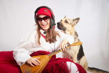 A girl in red scarf, white shirt, with shawl and balalaika in black sunglasses with a shepherd dog....